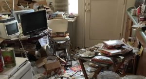 A cluttered room with various items scattered around, including a TV, microwave, boxes, papers, and food containers. The floor is covered with debris, and there is an air conditioner in the window.
