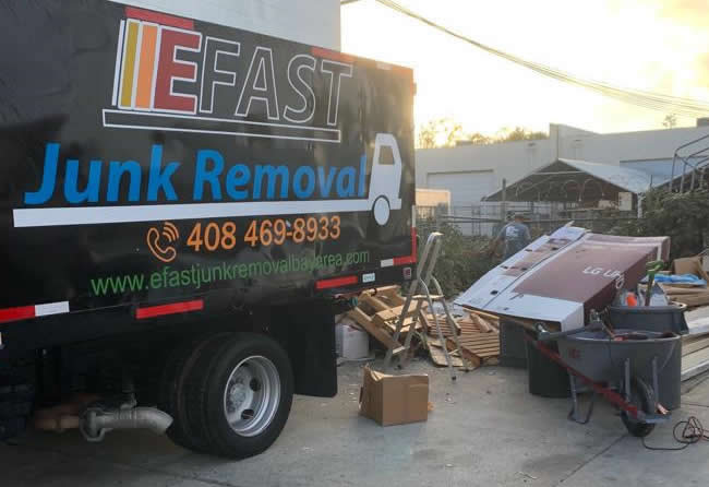 A black truck labeled "EFast Junk Removal" stands next to a pile of discarded items, including a large broken sign and miscellaneous debris. The contact number and website are displayed on the truck's side.