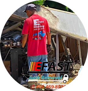 A man in a red shirt with "Fast Junk Removal" text, stands near a pile of clutter and debris next to a broken wooden structure. Contact number on shirt: (408) 469-8933.