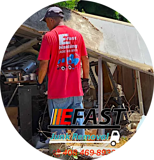 A man in a red shirt with "Fast Junk Removal" text, stands near a pile of clutter and debris next to a broken wooden structure. Contact number on shirt: (408) 469-8933.