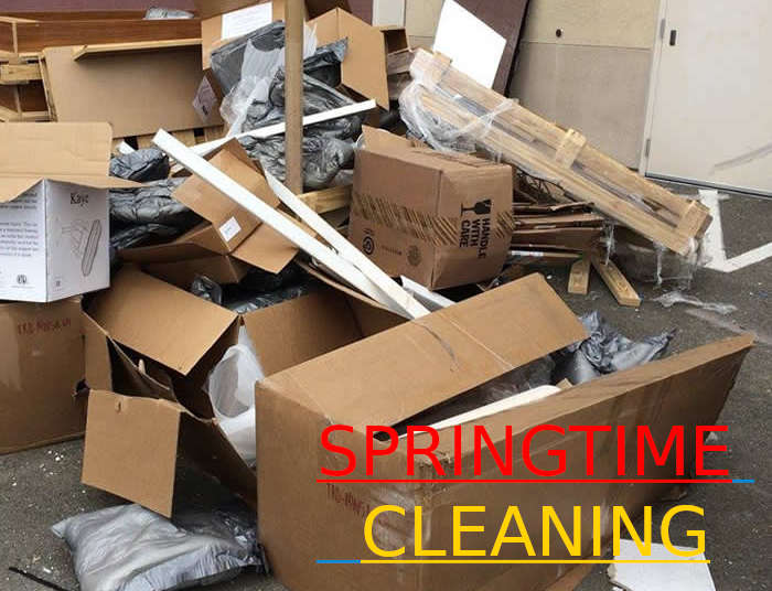 A pile of broken-down cardboard boxes, wooden panels, and plastic bags on the ground, with the text "Springtime Cleaning" in large red and yellow letters overlaying the image.
