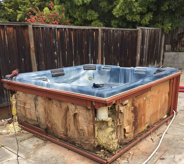 A blue hot tub with damaged and exposed insulation is located outdoors on a concrete surface, next to a wooden fence and surrounded by various tools and debris.