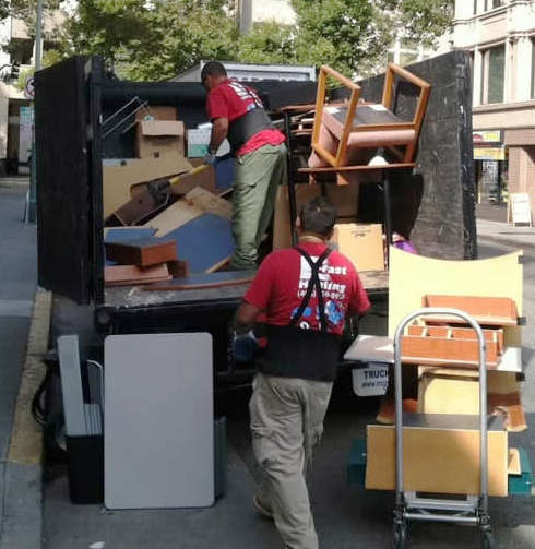 Two movers unload furniture from a large truck onto the street. The items include chairs, tables, and a rolling cart. The scene is set in an urban environment with buildings and trees in the background.