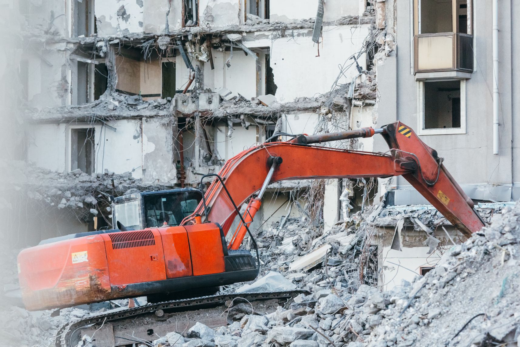 A red excavator demolishes a partially destroyed concrete building surrounded by rubble.