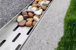 Metal drainage channel filled with various smooth, colorful stones, positioned between a gravel path and a grassy area.