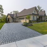 A single-story house with a large cobblestone driveway, manicured lawn, and surrounding trees seen on a clear day.