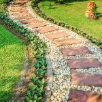 A curving garden path made of square stone slabs and white pebbles, lined with small plants on a neatly trimmed green lawn.