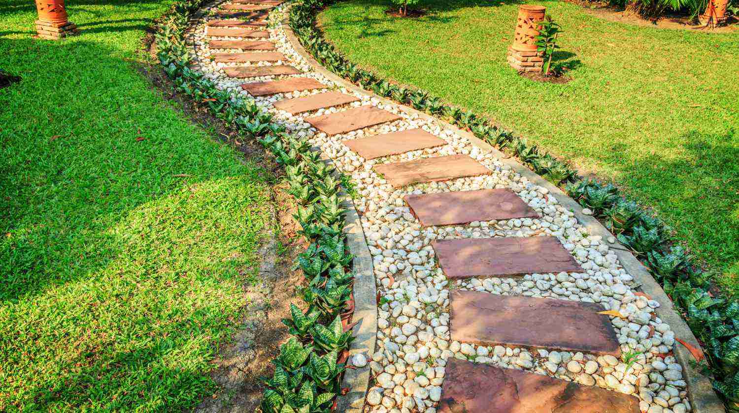 A curving garden path made of square stone slabs and white pebbles, lined with small plants on a neatly trimmed green lawn.