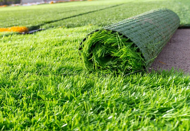 A roll of artificial grass partially unrolled on the ground, showing the green synthetic fibers and black backing material.