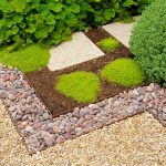 A garden section with green plants, square stepping stones, green moss, and multicolored pebbles arranged in neat patterns.
