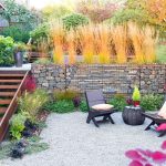 Outdoor garden area with two dark-colored chairs and a small table on gravel. Wooden stairs lead up to a higher level with ornamental grasses and potted plants. Shrubs and foliage surround the space.