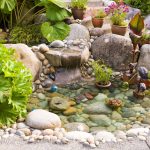 A small garden pond with clear water, surrounded by large stones and potted plants. The pond features a small waterfall, and various green and flowering plants are placed around it.