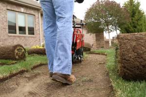 Person in blue jeans and brown boots operates a red sod cutter on a lawn, with rolled-up sod pieces visible.